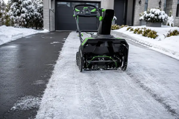 Black and green cordless snow blower on a driveway with a visible thin layer of snow left behind one clearing pass.